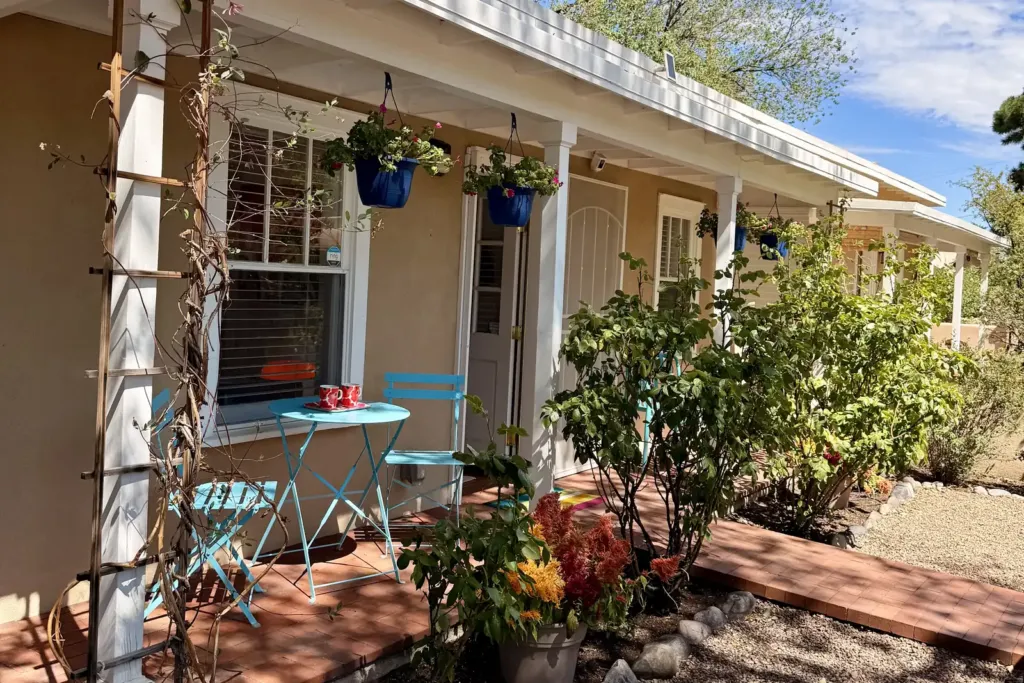 Front porch of a Santa Fe vacation rental casita with blue bistro chairs, hanging flower baskets and a gravel garden